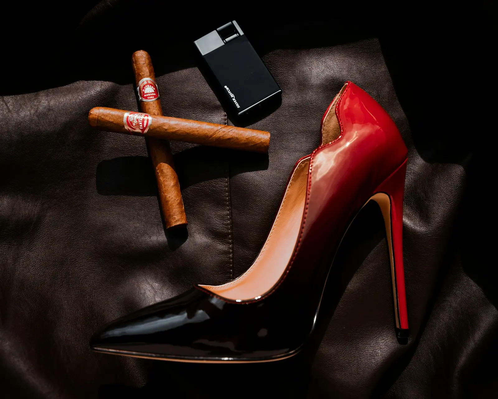 Stiletto rojo de charol junto a dos habanos sobre cuero negro